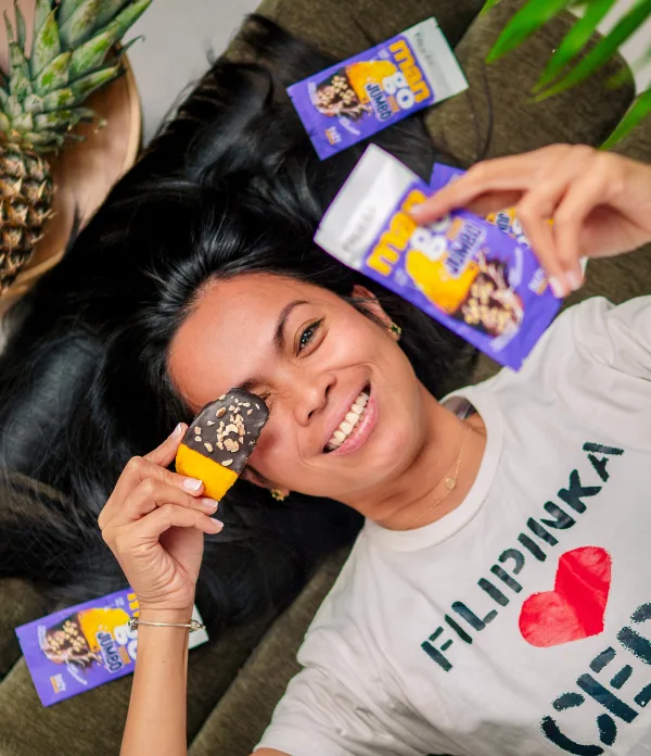Uśmiechnięta kobieta leżąca z produktami ManGo Jumbo. Trzyma plaster suszonego mango w czekoladzie, zasłaniając nim jedno oko.